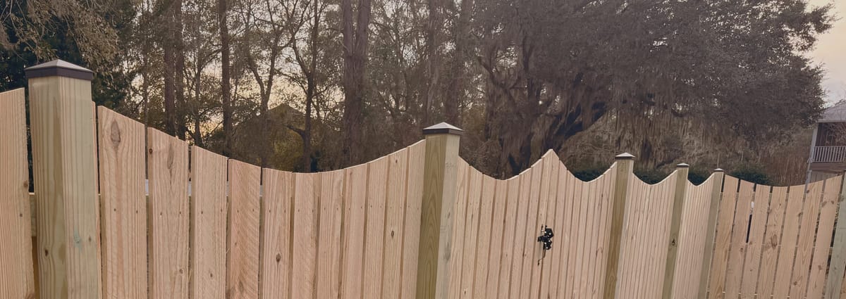 A finished wood privacy fence with a scalloped top line and black post caps running away under live oaks hung with Spanish moss.