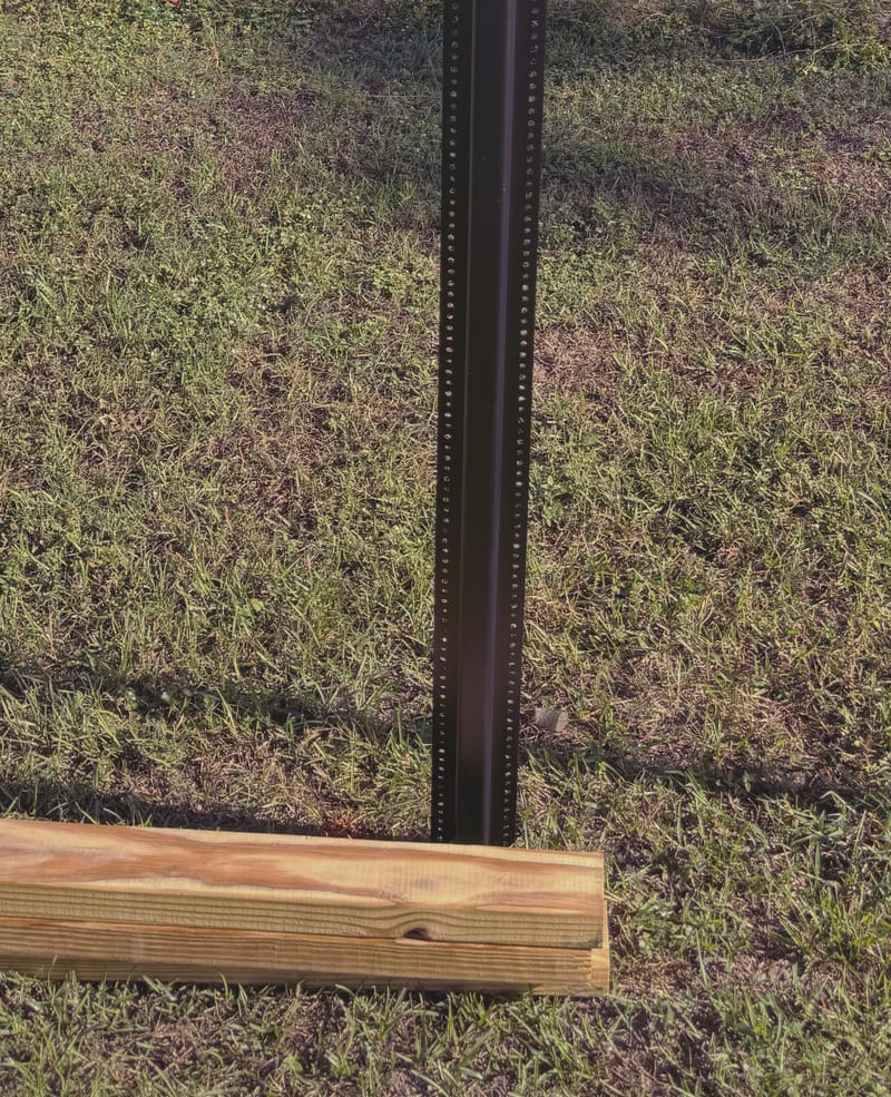 A single black steel post with rows of punched holes standing upright in a lawn, with a fresh timber rail lying on the grass beside it.