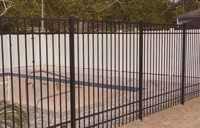 A black aluminum pool fence with a self closing gate enclosing a swimming pool, with a privacy fence behind it.